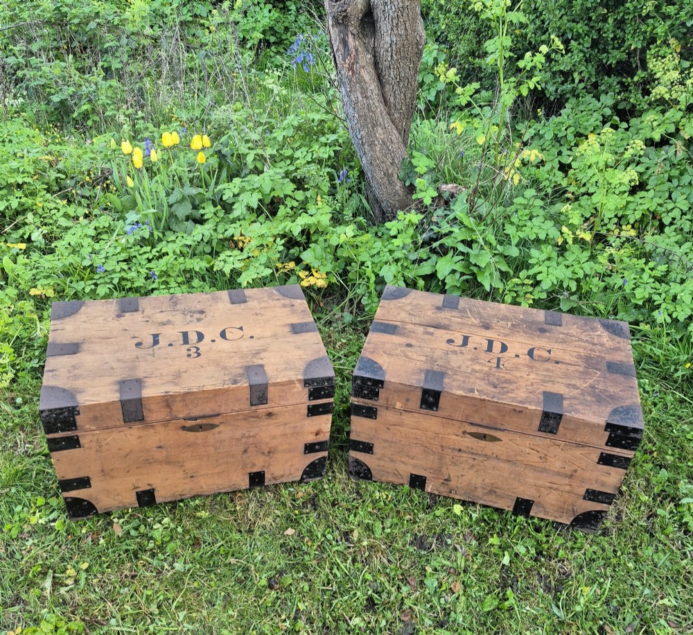 A pair of rectangular natural pine chests featuring black iron corner brackets, metal straps, keyhole escutcheons, and black stencilled text on the lids.