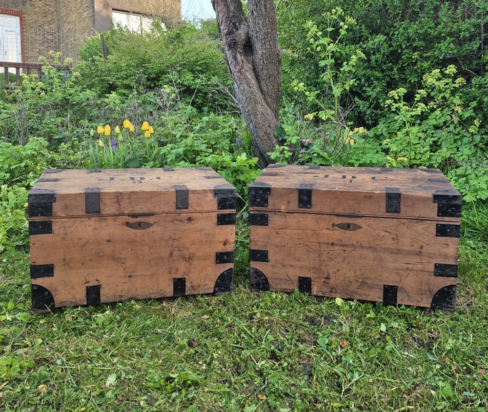 A pair of rectangular natural pine chests featuring black iron corner brackets, metal straps, keyhole escutcheons, and black stencilled text on the lids. View II