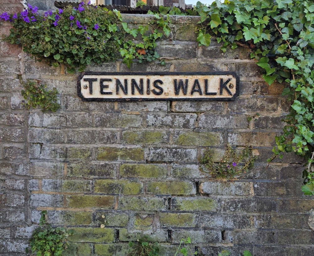 A rectangular 1920s cast iron street sign featuring embossed black lettering spelling TENNIS WALK on a weathered off-white background with a black border. View VII