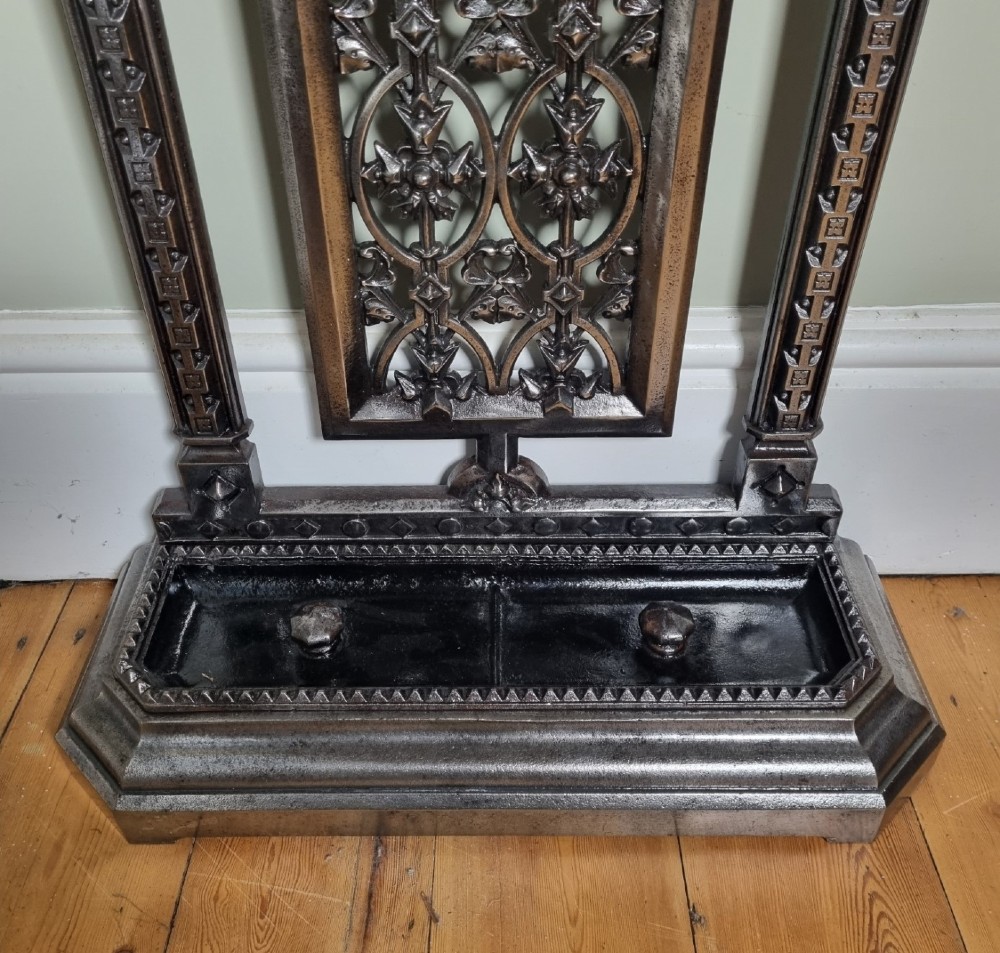 A Victorian cast iron stick stand featuring a pierced Gothic arch design, retention bar, and a rectangular drip tray base. View V
