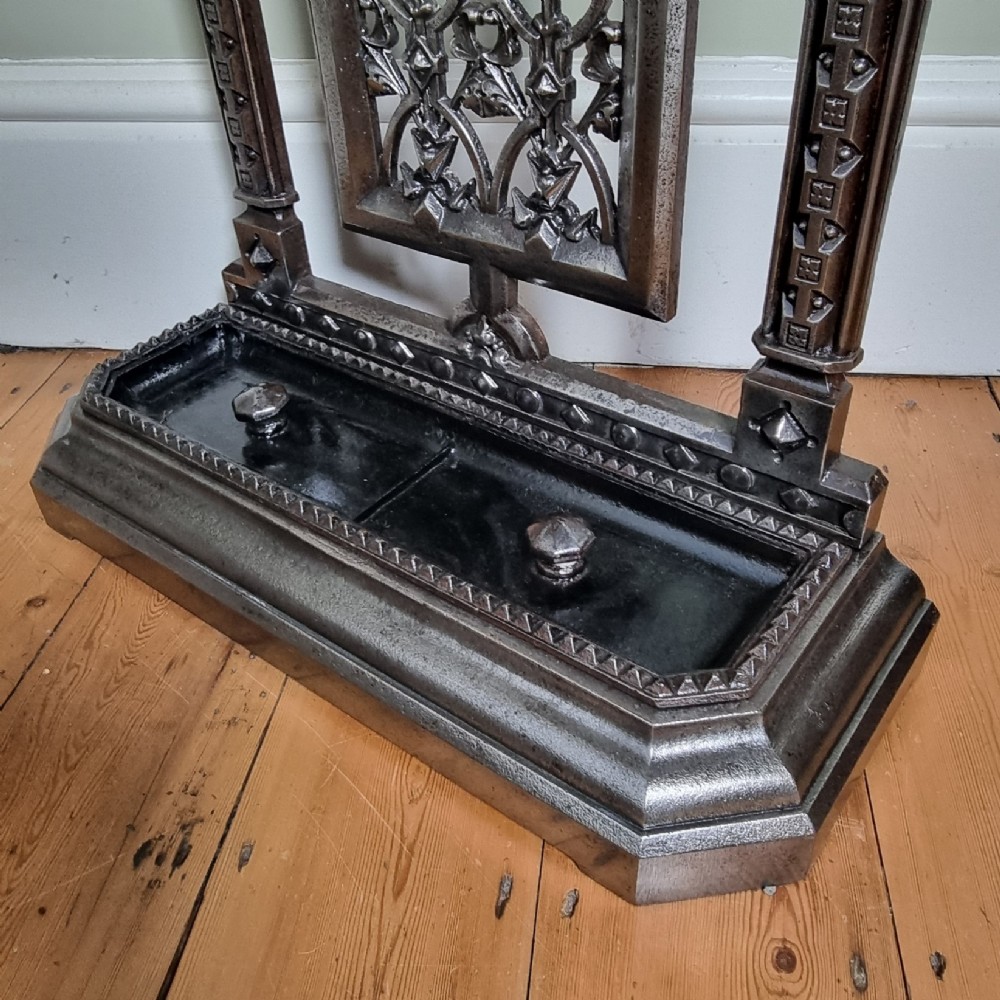 A Victorian cast iron stick stand featuring a pierced Gothic arch design, retention bar, and a rectangular drip tray base. View VI