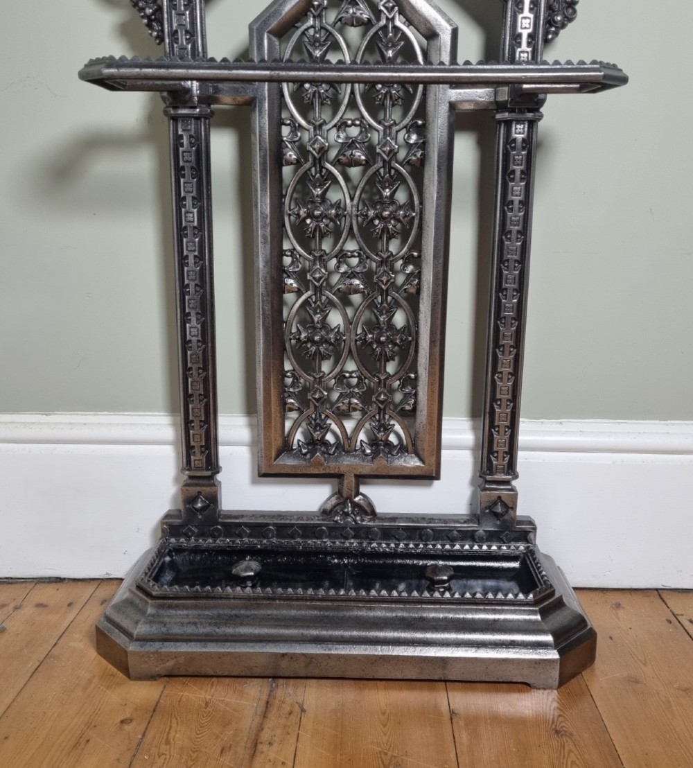 A Victorian cast iron stick stand featuring a pierced Gothic arch design, retention bar, and a rectangular drip tray base. View X