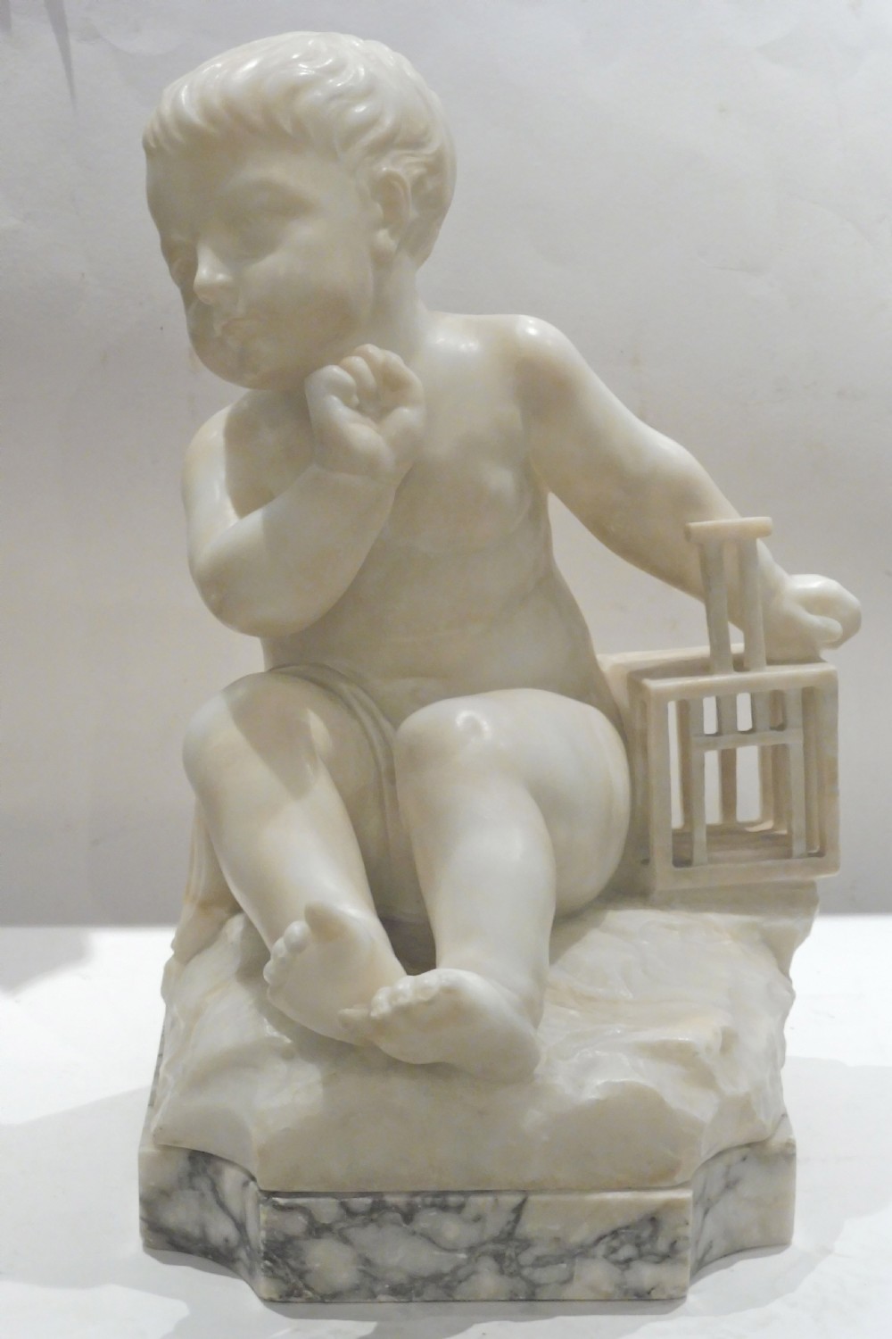An Italian carved alabaster sculpture depicting a young seated boy beside a miniature birdcage, set on a grey veined marble base.