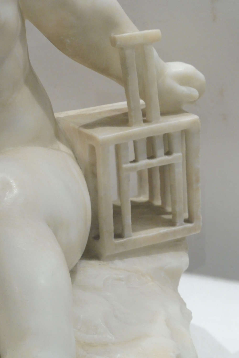 An Italian carved alabaster sculpture depicting a young seated boy beside a miniature birdcage, set on a grey veined marble base. View III