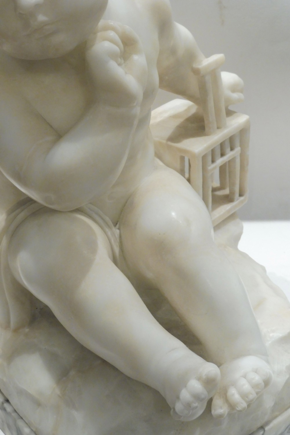 An Italian carved alabaster sculpture depicting a young seated boy beside a miniature birdcage, set on a grey veined marble base. View IV