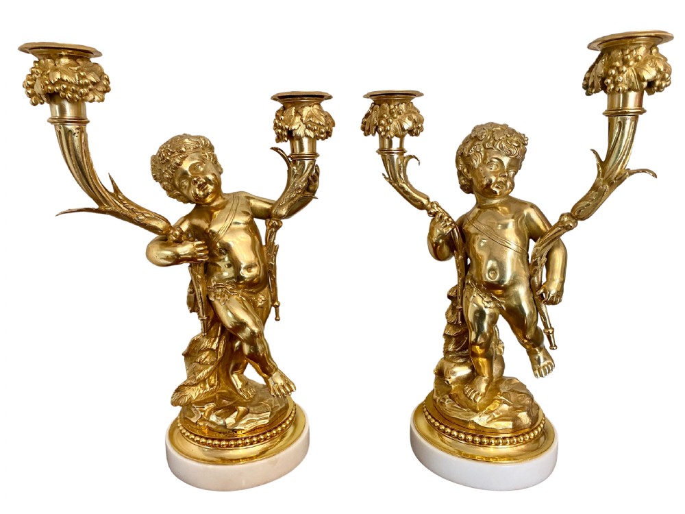 A pair of gilt bronze two-arm candelabra featuring sculpted cherub figures resting on circular white marble bases.