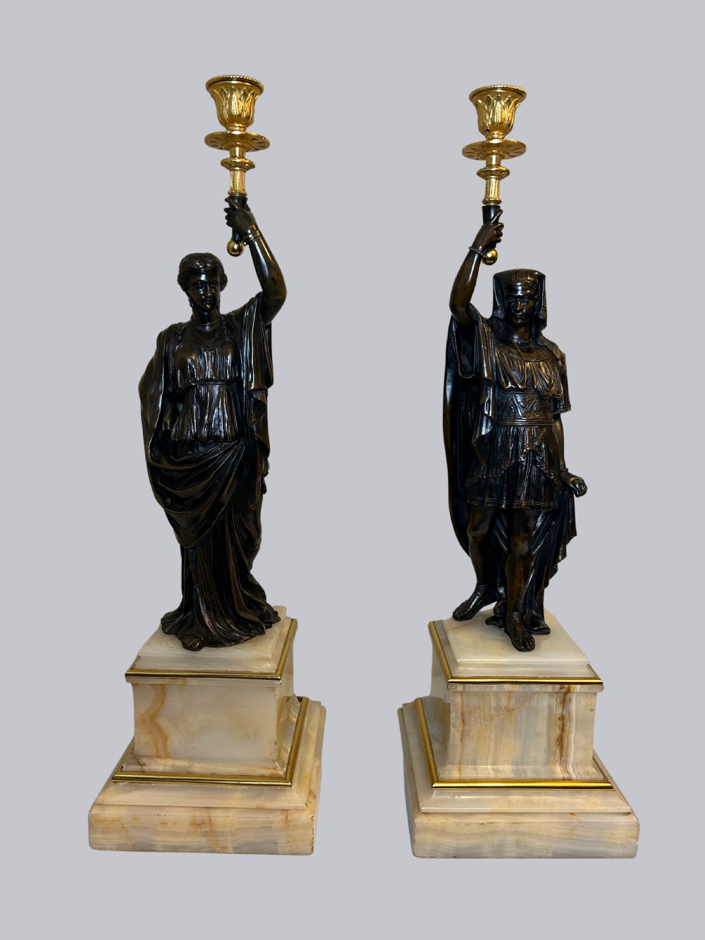 A pair of 19th-century French patinated bronze figural candlesticks featuring draped male and female classical statues holding gilt sconces atop stepped cream onyx bases.