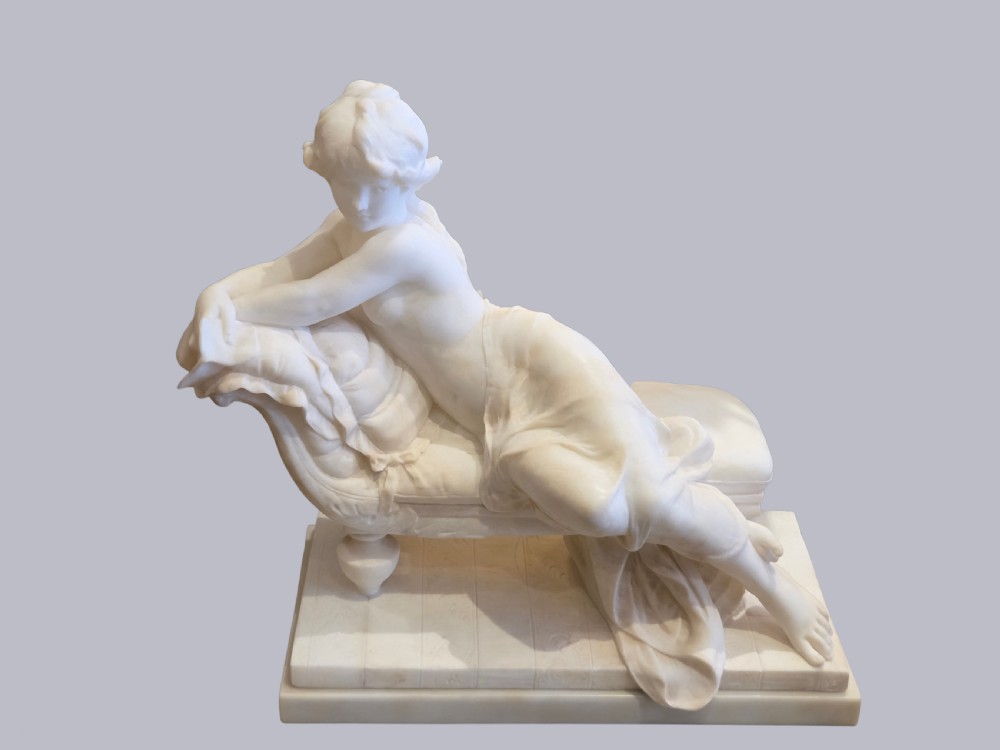 A carved off-white alabaster sculpture depicting a semi-clad reclining woman resting on a chaise longue set on a rectangular plinth.