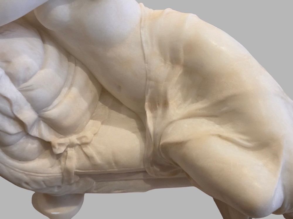 A carved off-white alabaster sculpture depicting a semi-clad reclining woman resting on a chaise longue set on a rectangular plinth. View VI