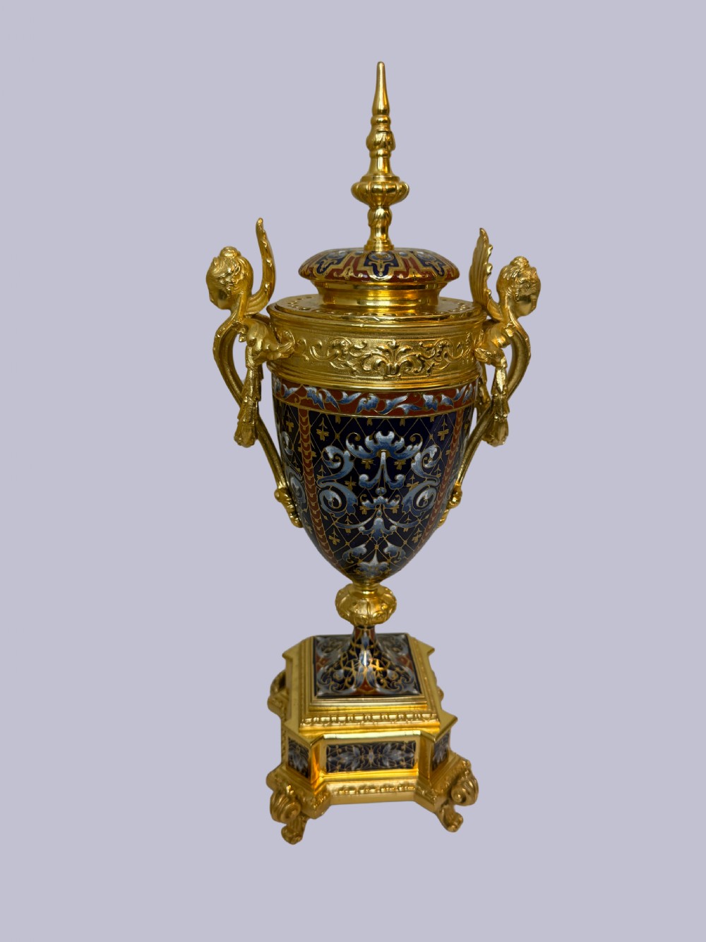 A 19th-century French lidded urn crafted from gilt bronze featuring blue and red champleve enamel detailing with figural handles and a finial lid.