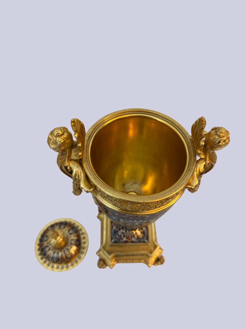 A 19th-century French lidded urn crafted from gilt bronze featuring blue and red champleve enamel detailing with figural handles and a finial lid. View V