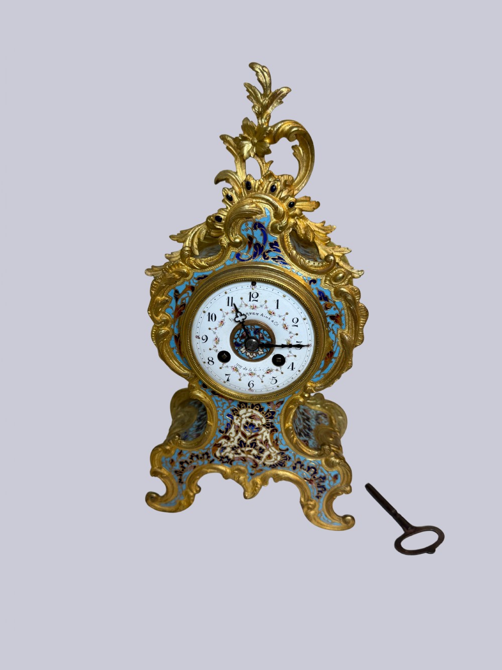 An ornate French Rococo Revival mantel clock featuring gilt bronze scrollwork, light blue champleve enamel patterns, and a white enamel dial with floral decoration, shown with a winding key.