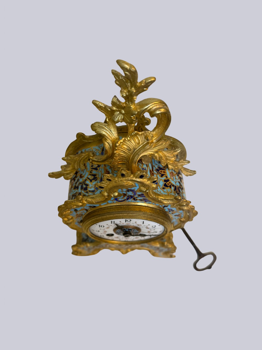 An ornate French Rococo Revival mantel clock featuring gilt bronze scrollwork, light blue champleve enamel patterns, and a white enamel dial with floral decoration, shown with a winding key. View II