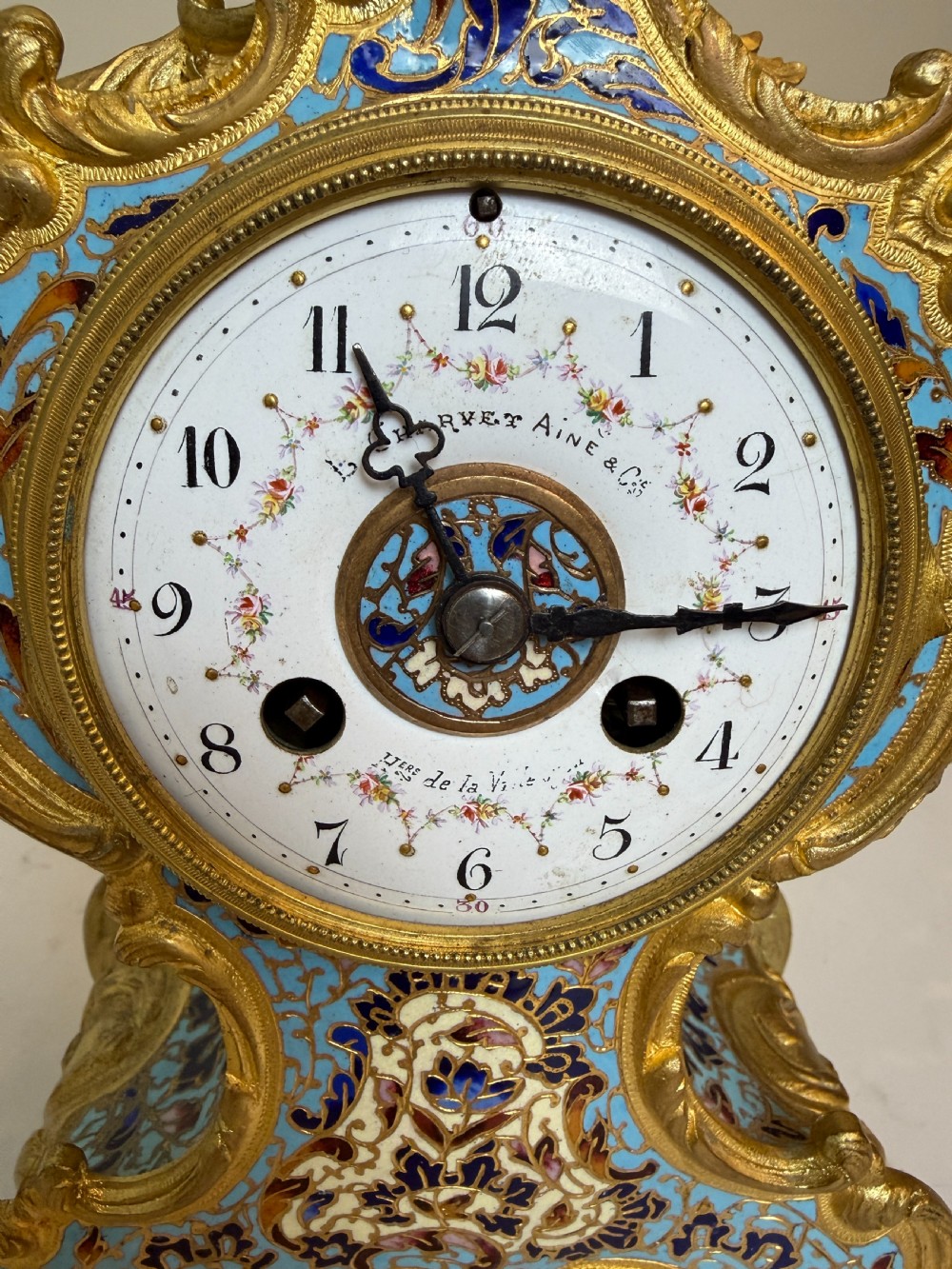 An ornate French Rococo Revival mantel clock featuring gilt bronze scrollwork, light blue champleve enamel patterns, and a white enamel dial with floral decoration, shown with a winding key. View IV