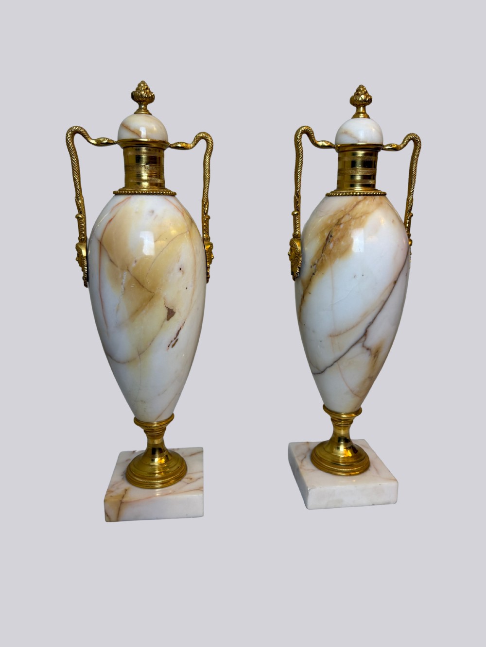 A pair of 19th-century ovoid white onyx urns featuring gilt bronze serpent handles, finials, and square pedestals.