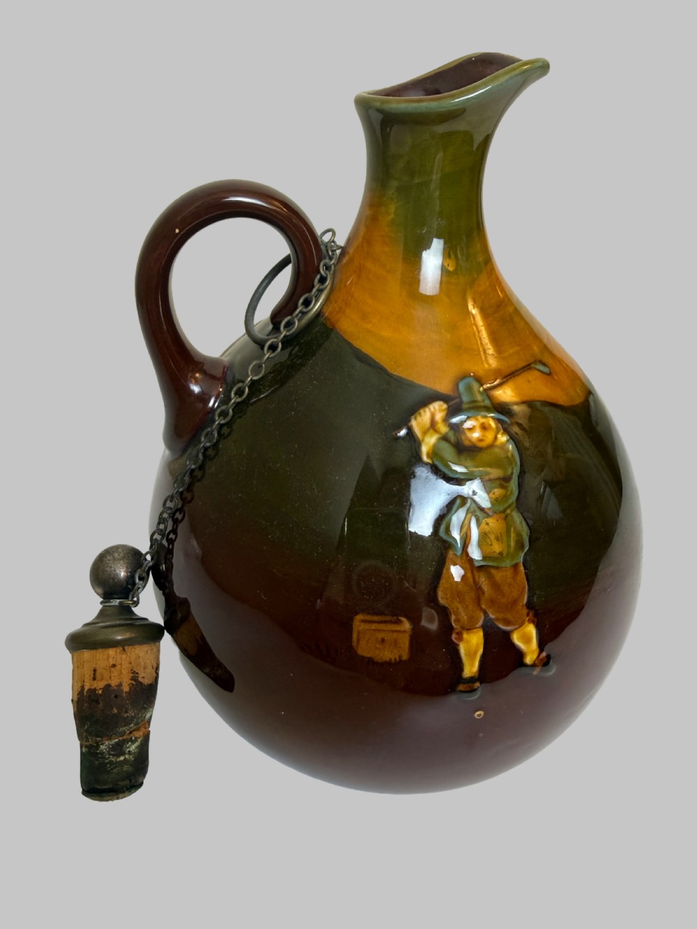 A brown and ochre glazed ceramic whisky flask featuring a raised relief figure of a golfer with an attached cork stopper on a metal chain.