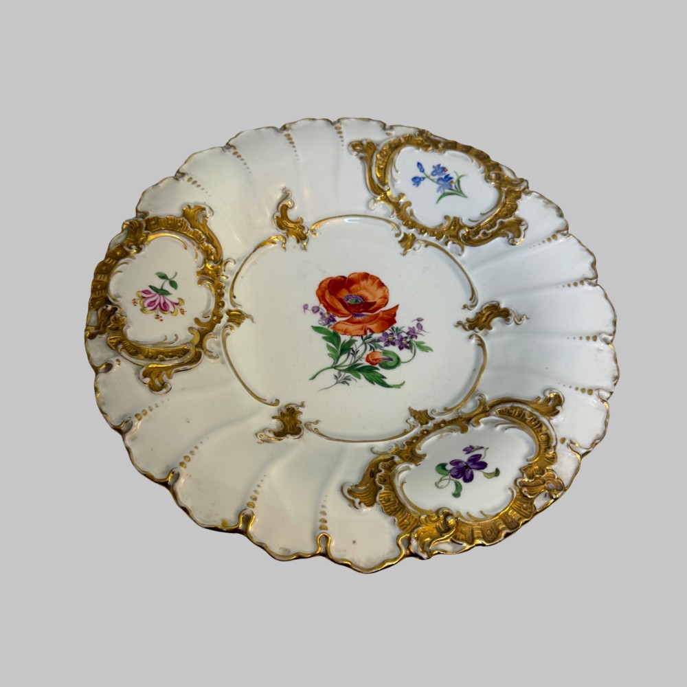 A white scalloped porcelain dish featuring hand-painted central botanical flowers surrounded by gilded Rococo cartouches containing smaller floral sprays.