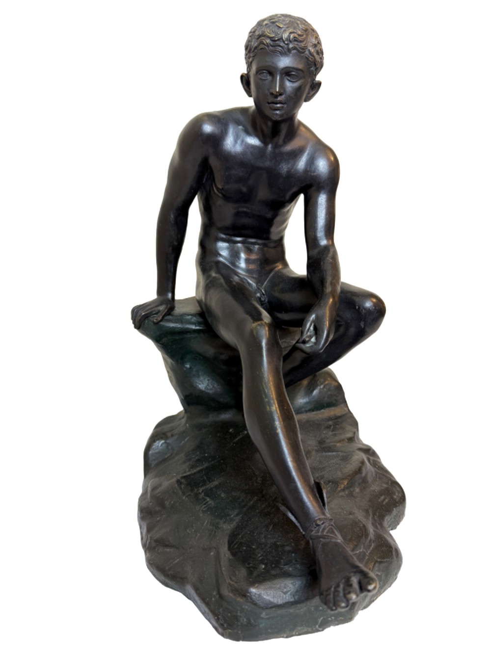 A dark patinated bronze sculpture depicting a classical male nude youth resting seated upon a rugged stone formation.