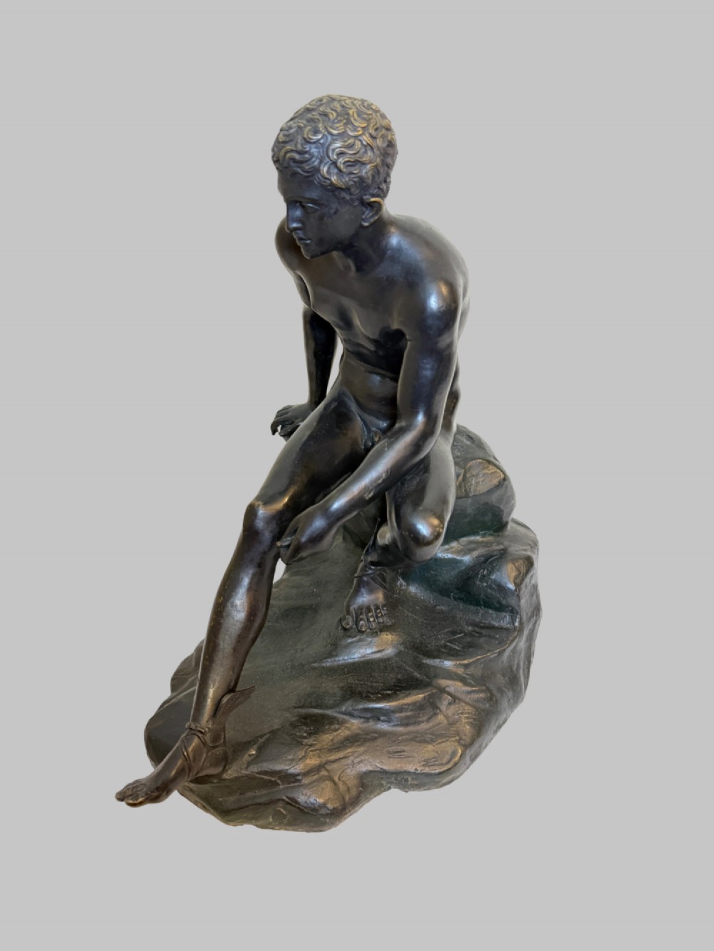 A dark patinated bronze sculpture depicting a classical male nude youth resting seated upon a rugged stone formation. View II