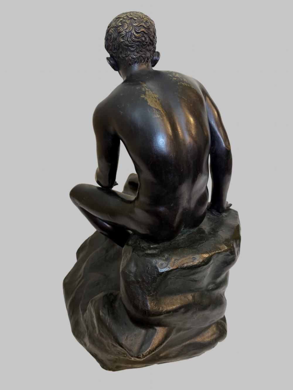 A dark patinated bronze sculpture depicting a classical male nude youth resting seated upon a rugged stone formation. View III