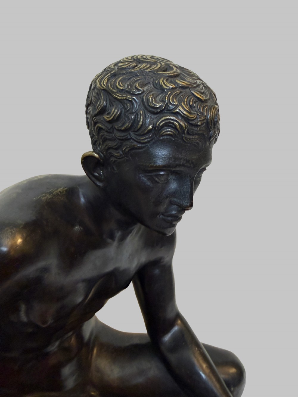 A dark patinated bronze sculpture depicting a classical male nude youth resting seated upon a rugged stone formation. View V