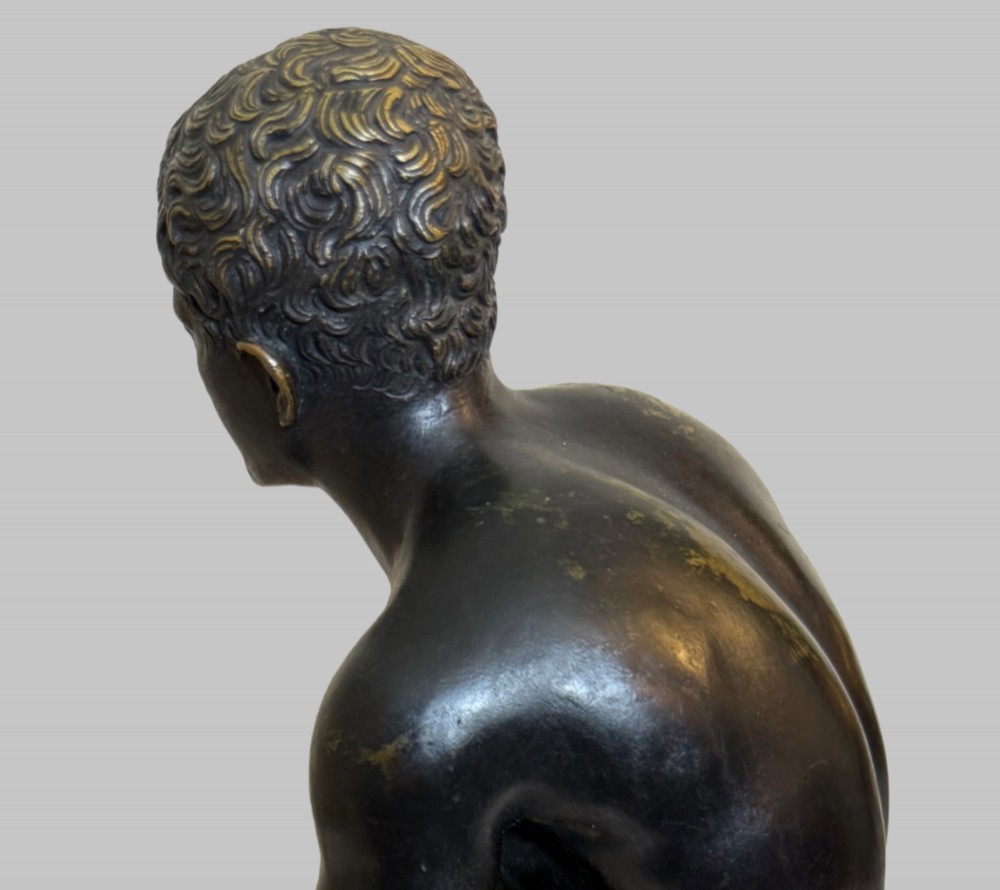 A dark patinated bronze sculpture depicting a classical male nude youth resting seated upon a rugged stone formation. View VII