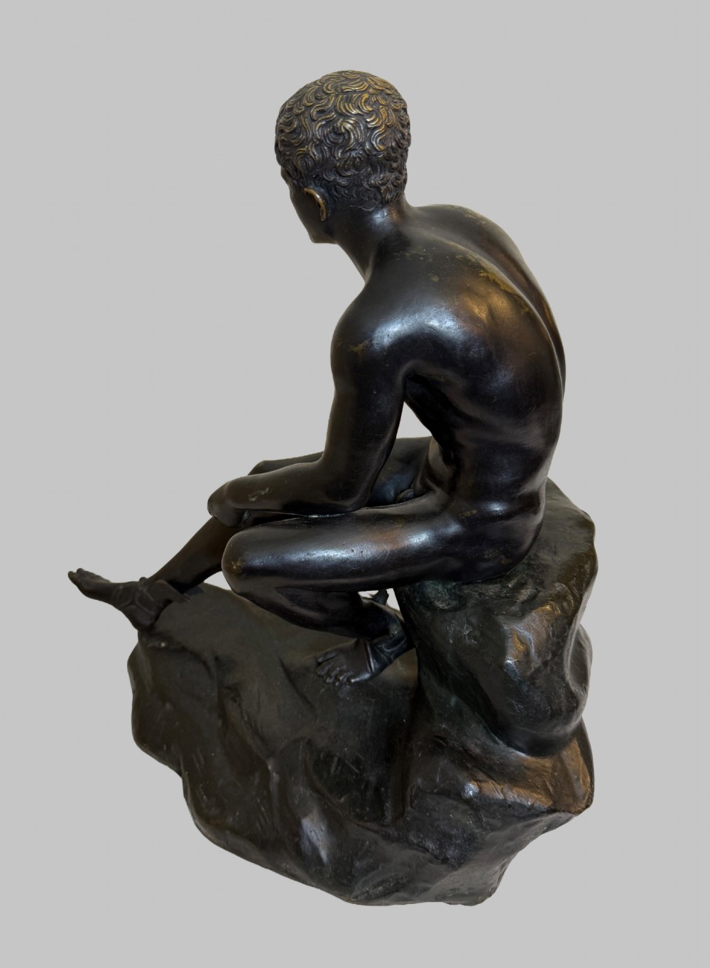 A dark patinated bronze sculpture depicting a classical male nude youth resting seated upon a rugged stone formation. View VIII