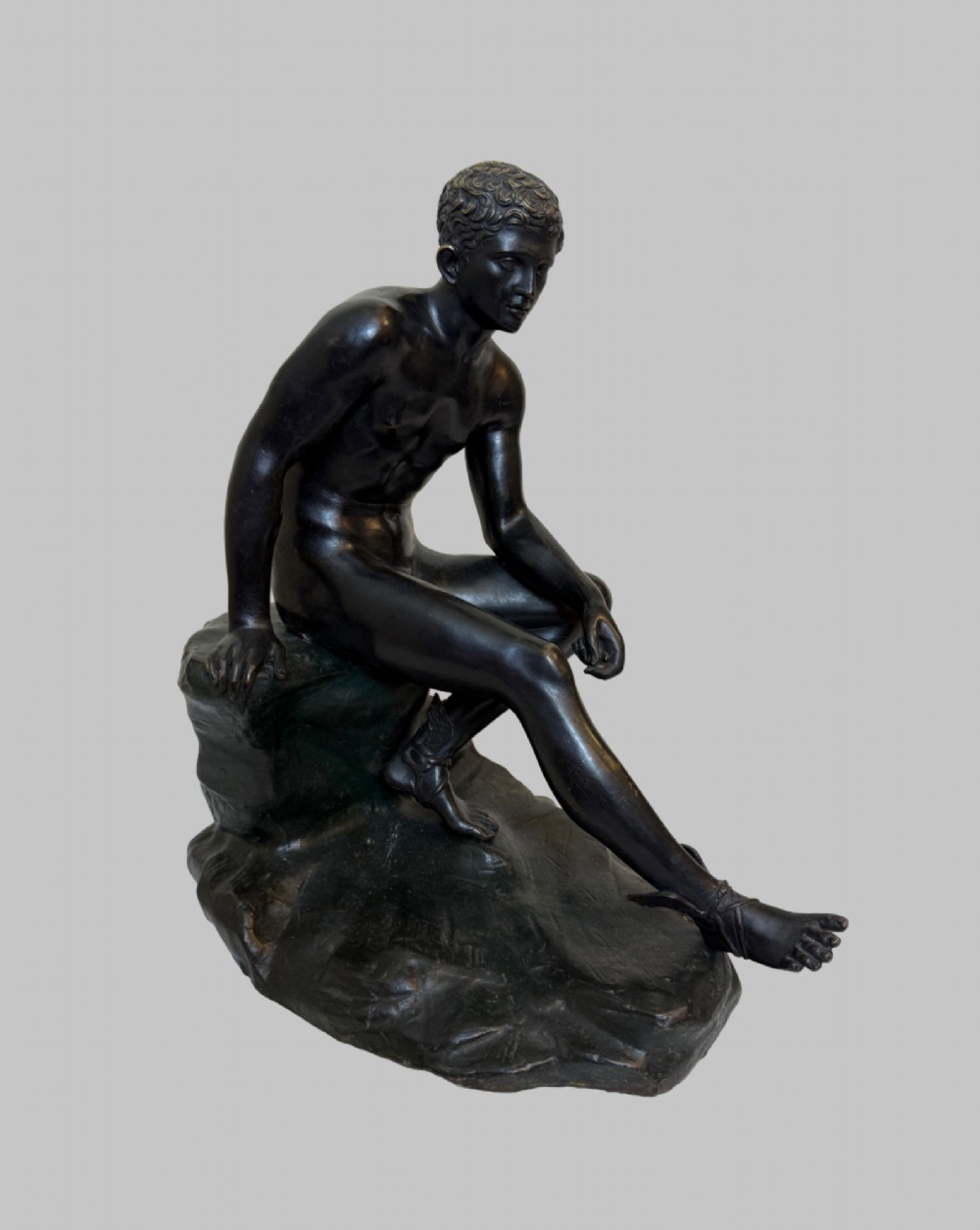 A dark patinated bronze sculpture depicting a classical male nude youth resting seated upon a rugged stone formation. View IX