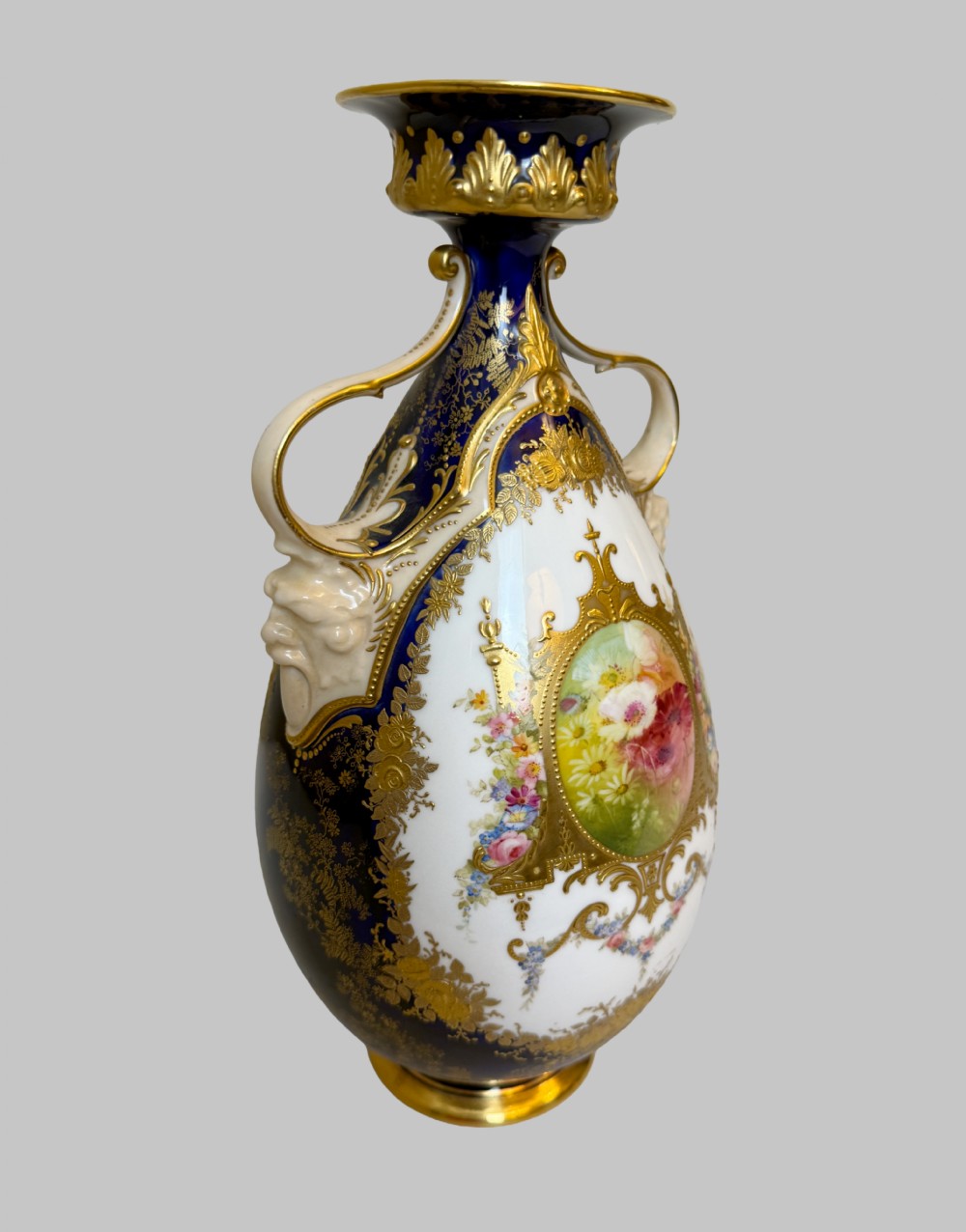 An antique Royal Doulton porcelain vase featuring cobalt blue and white glazes, heavy gilt scrolling, scroll handles with mask terminals, and a central hand-painted floral panel. View II