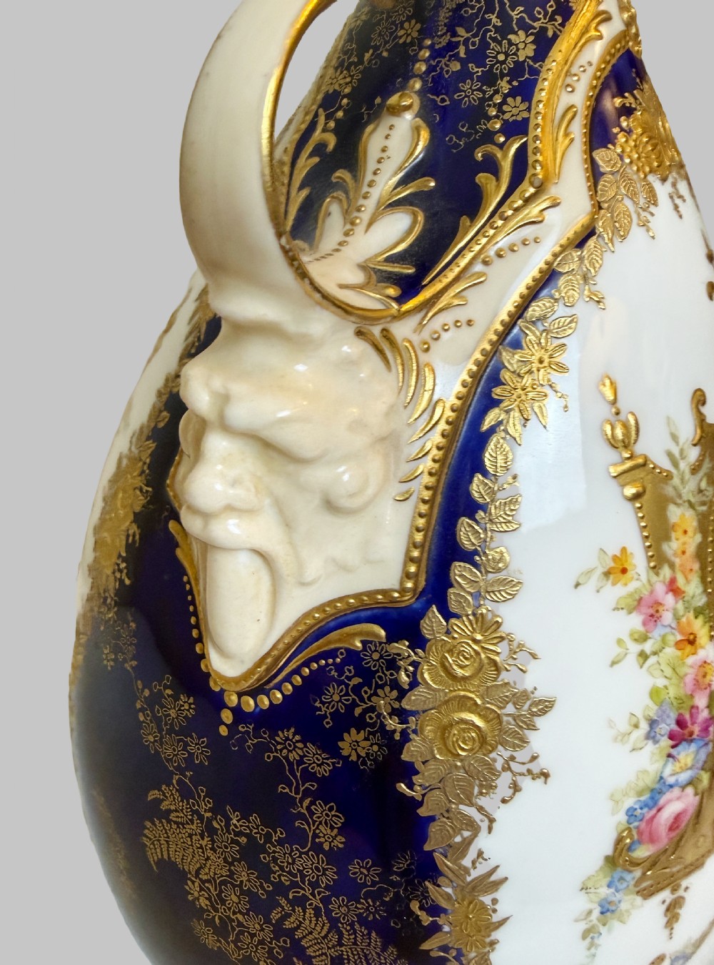 An antique Royal Doulton porcelain vase featuring cobalt blue and white glazes, heavy gilt scrolling, scroll handles with mask terminals, and a central hand-painted floral panel. View IV