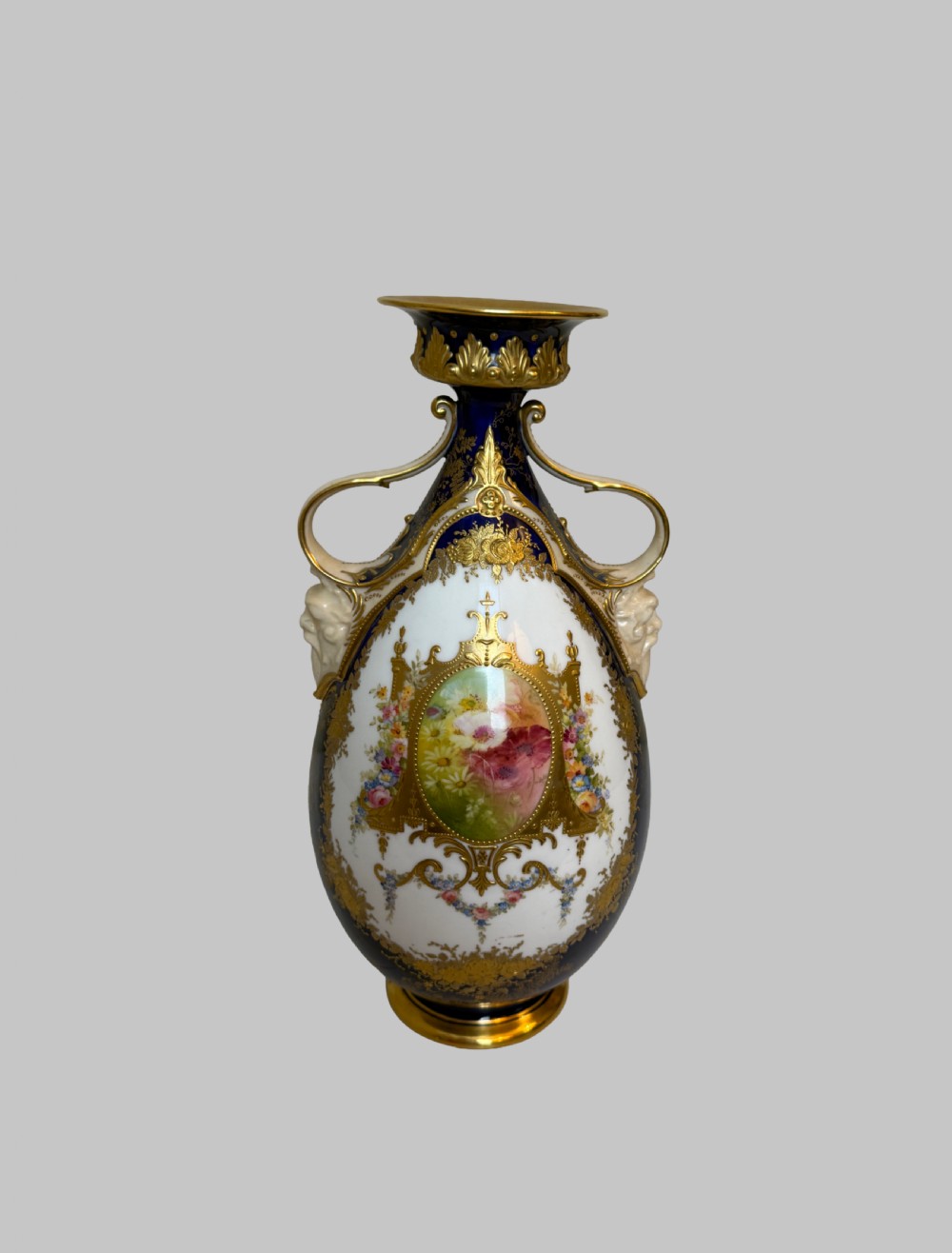 An antique Royal Doulton porcelain vase featuring cobalt blue and white glazes, heavy gilt scrolling, scroll handles with mask terminals, and a central hand-painted floral panel. View X