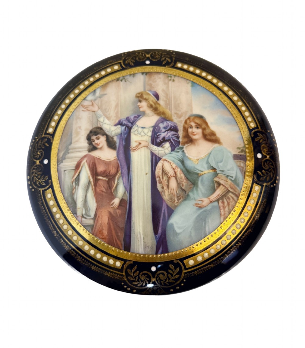 A circular antique French porcelain box featuring a central hand-painted classical scene of three women framed by cobalt blue, gilt embellishments, and white enamel dots.