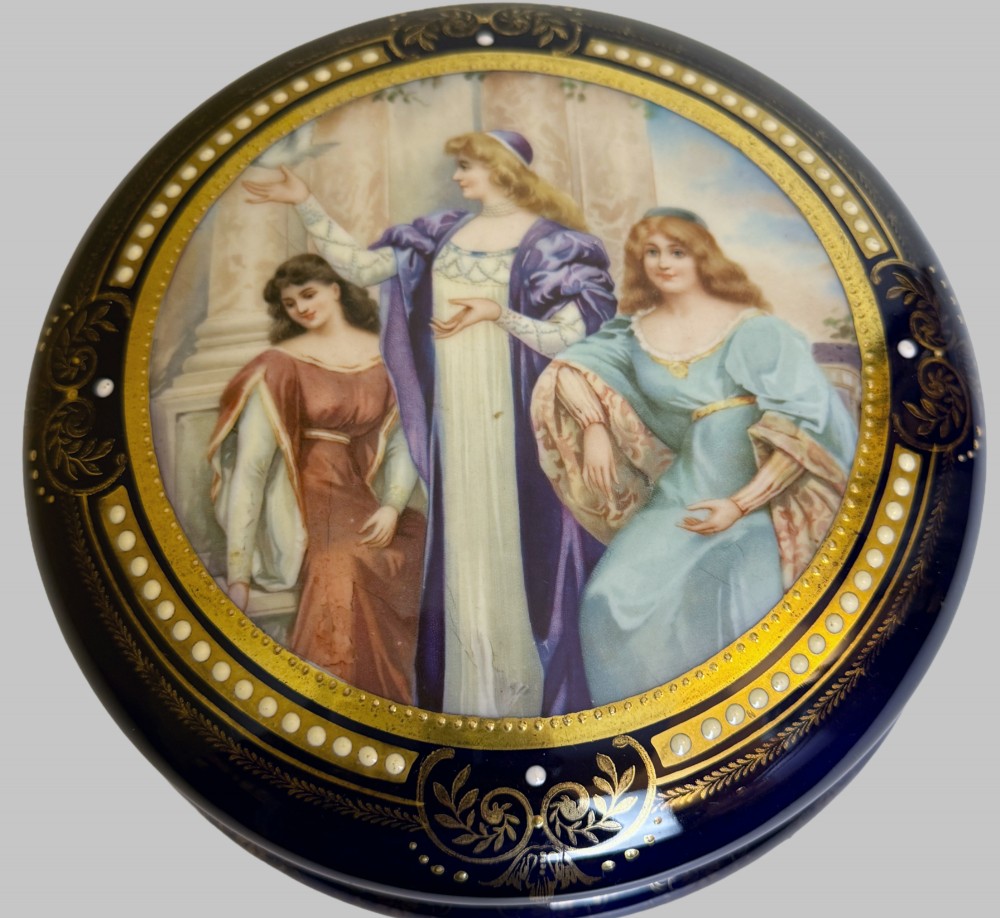 A circular antique French porcelain box featuring a central hand-painted classical scene of three women framed by cobalt blue, gilt embellishments, and white enamel dots. View II