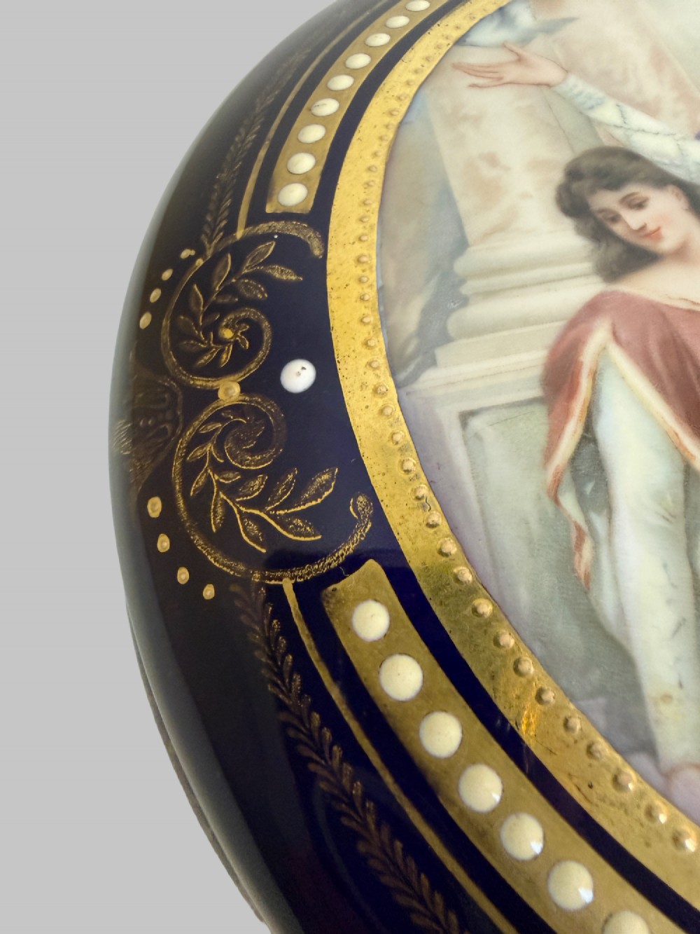 A circular antique French porcelain box featuring a central hand-painted classical scene of three women framed by cobalt blue, gilt embellishments, and white enamel dots. View III