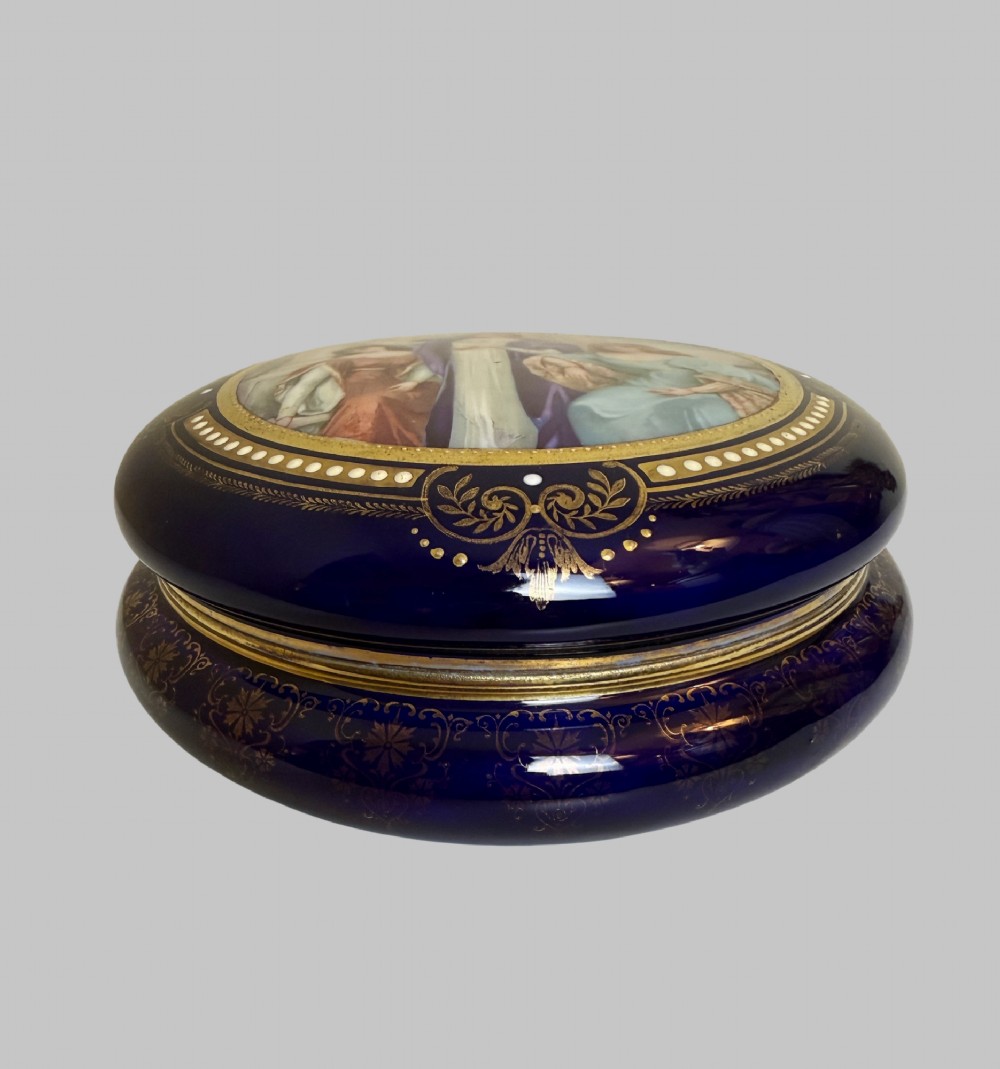A circular antique French porcelain box featuring a central hand-painted classical scene of three women framed by cobalt blue, gilt embellishments, and white enamel dots. View IV