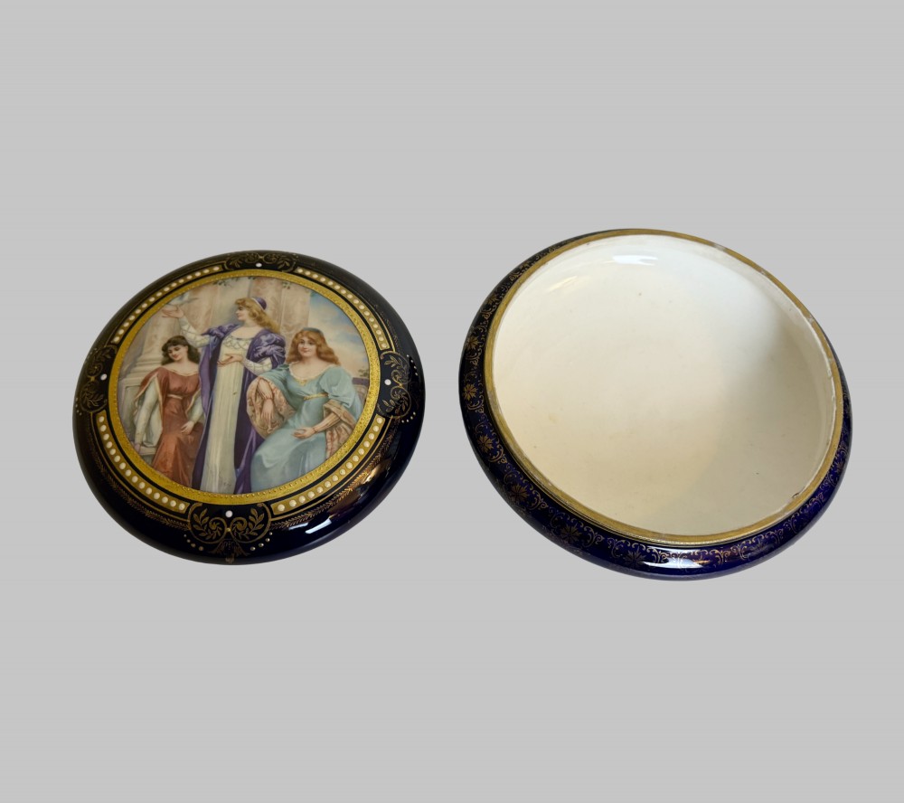 A circular antique French porcelain box featuring a central hand-painted classical scene of three women framed by cobalt blue, gilt embellishments, and white enamel dots. View V