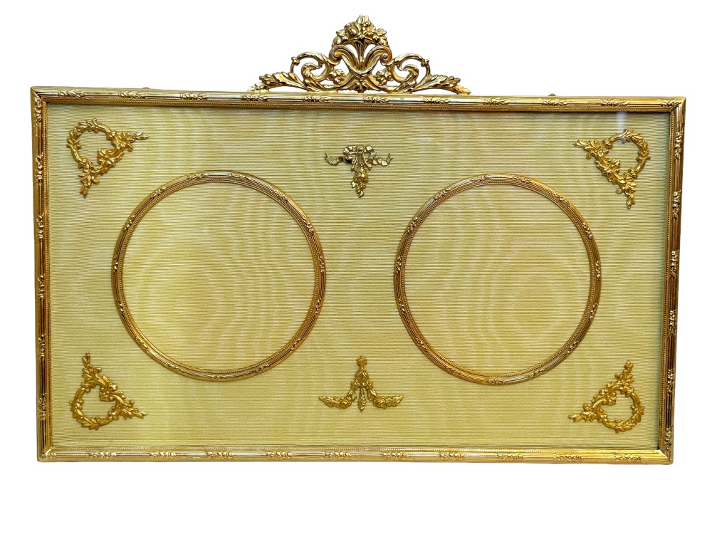 A 19th-century rectangular French gilt bronze frame featuring two circular photograph openings set against a cream silk background, adorned with a floral crest and ribbon mounts.