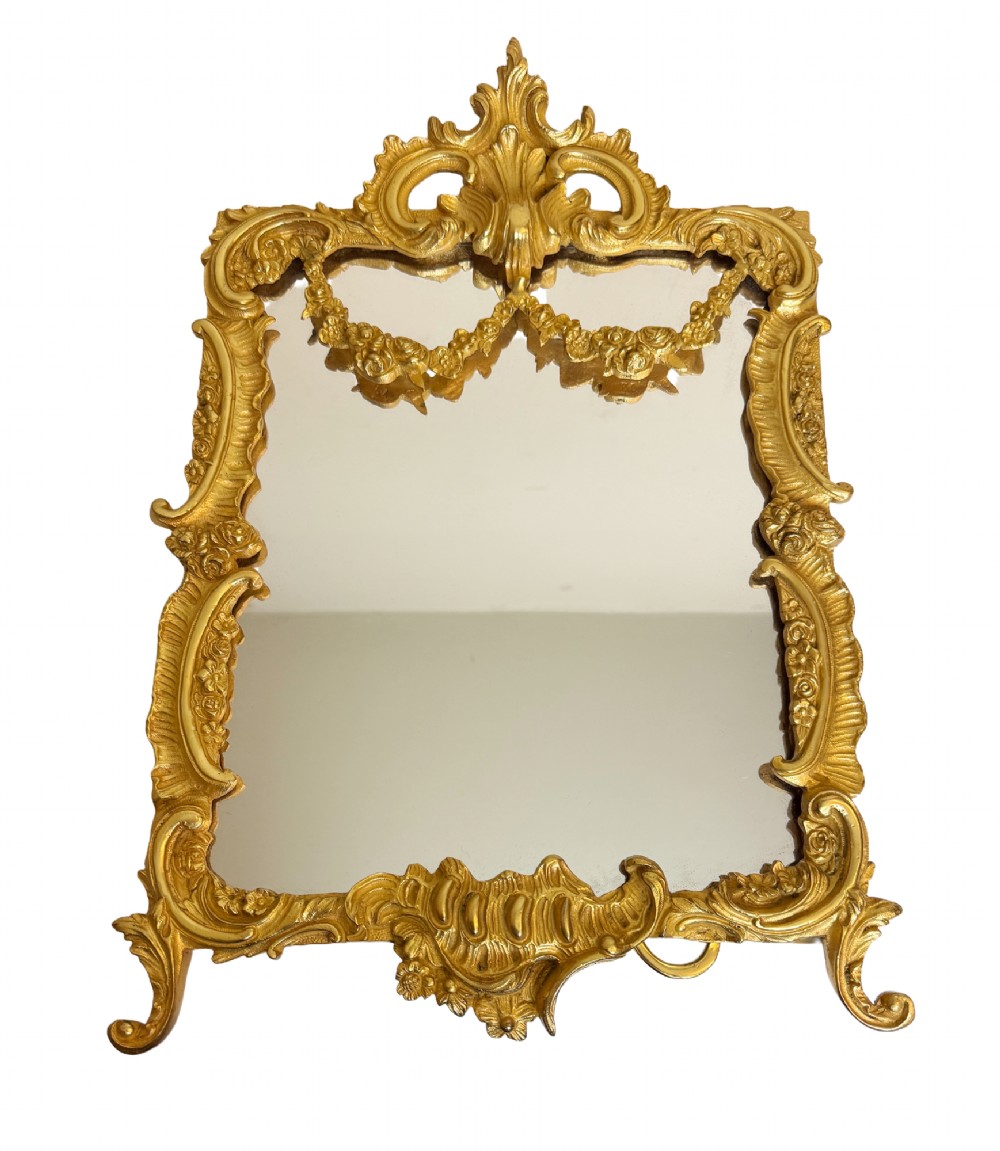 An ornate gold gilt bronze table mirror featuring Rococo scrollwork, floral garlands, and a carved crest motif.