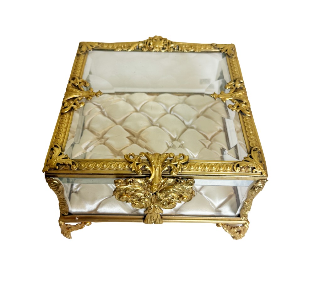 An antique rectangular bevelled crystal glass jewellery box featuring an ornate gilt bronze frame with decorative corner mounts and a button-tufted silk cushion interior.