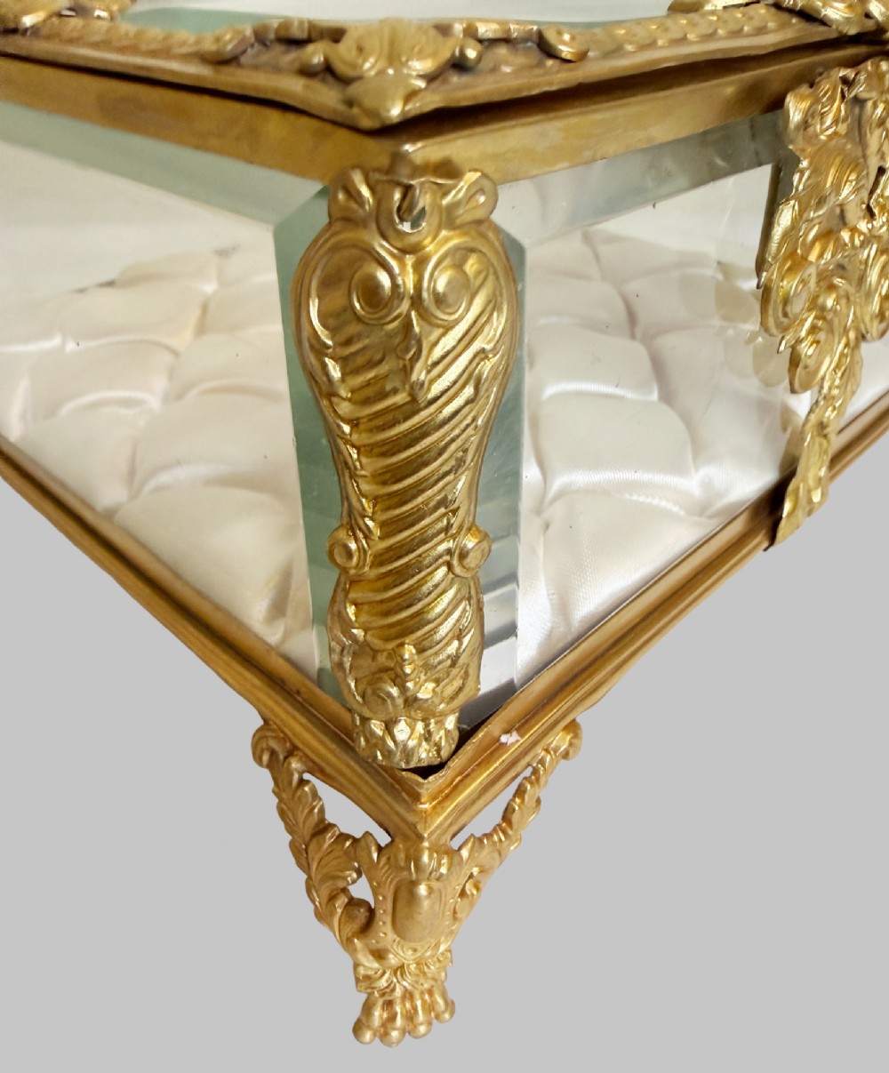 An antique rectangular bevelled crystal glass jewellery box featuring an ornate gilt bronze frame with decorative corner mounts and a button-tufted silk cushion interior. View V
