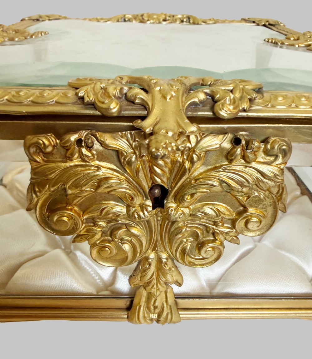 An antique rectangular bevelled crystal glass jewellery box featuring an ornate gilt bronze frame with decorative corner mounts and a button-tufted silk cushion interior. View VI