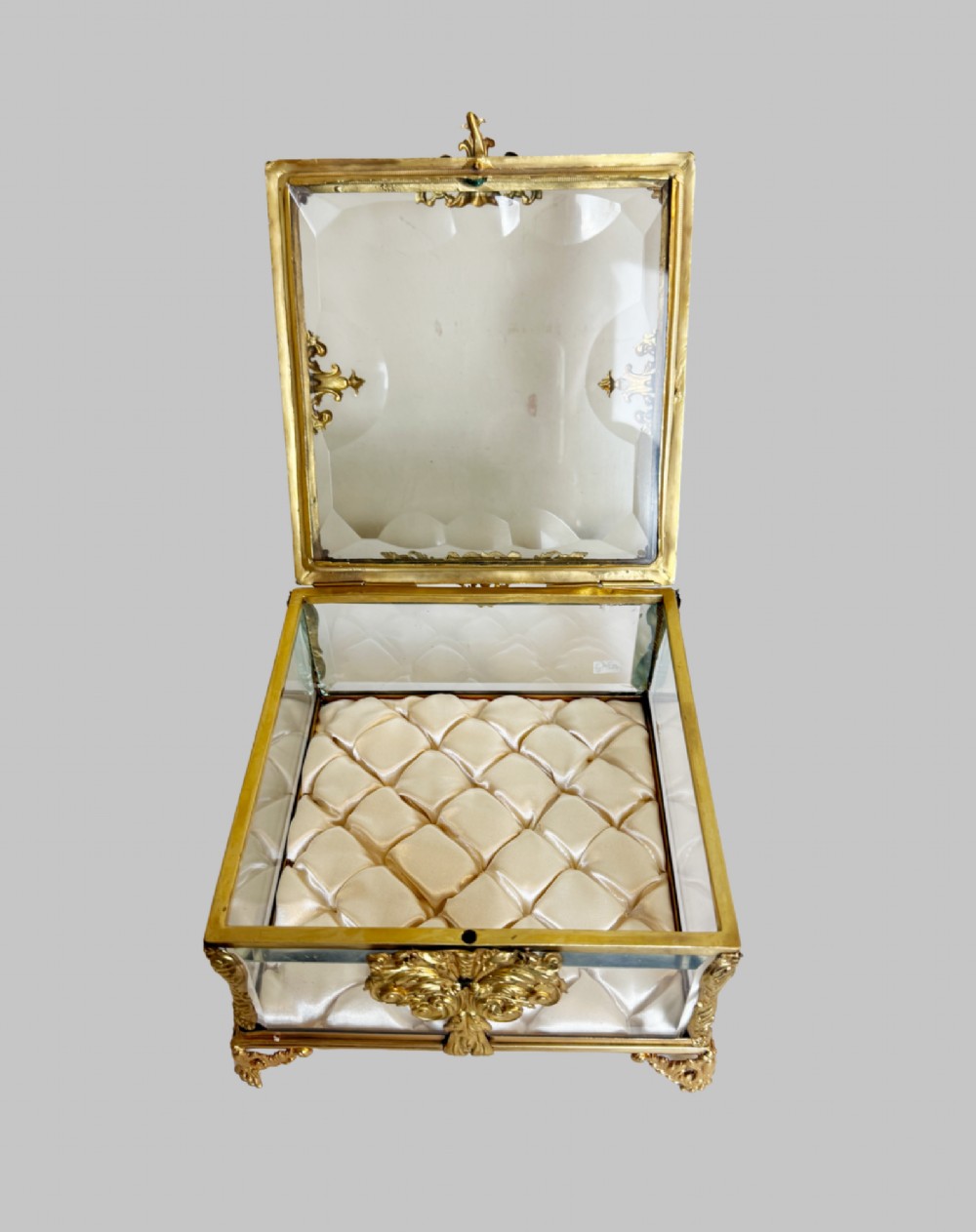 An antique rectangular bevelled crystal glass jewellery box featuring an ornate gilt bronze frame with decorative corner mounts and a button-tufted silk cushion interior. View VIII