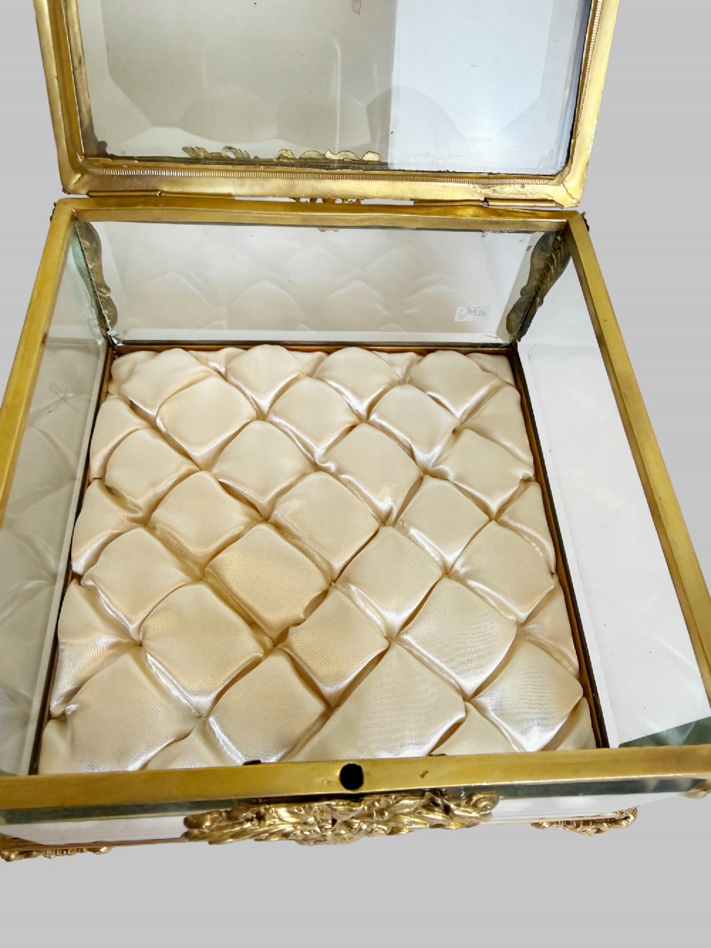 An antique rectangular bevelled crystal glass jewellery box featuring an ornate gilt bronze frame with decorative corner mounts and a button-tufted silk cushion interior. View IX