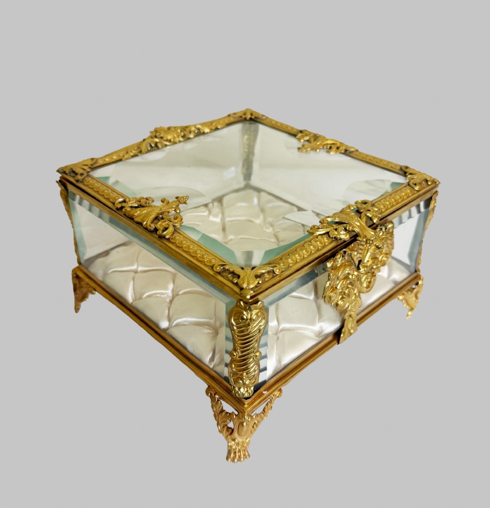 An antique rectangular bevelled crystal glass jewellery box featuring an ornate gilt bronze frame with decorative corner mounts and a button-tufted silk cushion interior. View X