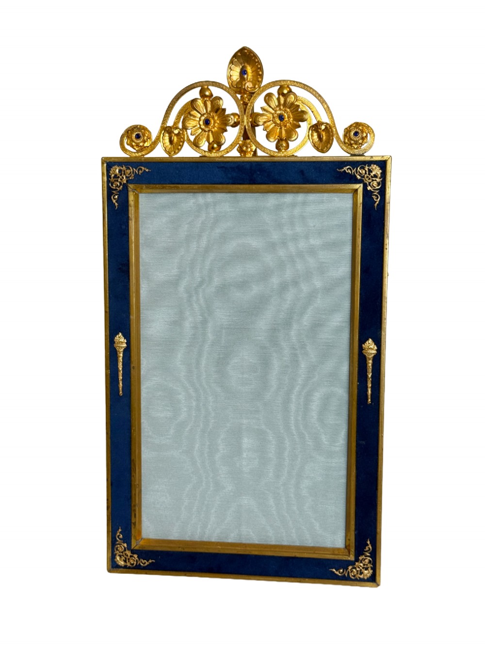 A rectangular gilt bronze picture frame featuring a blue velvet border, ornate corner mounts, and an elaborate floral crest at the top.