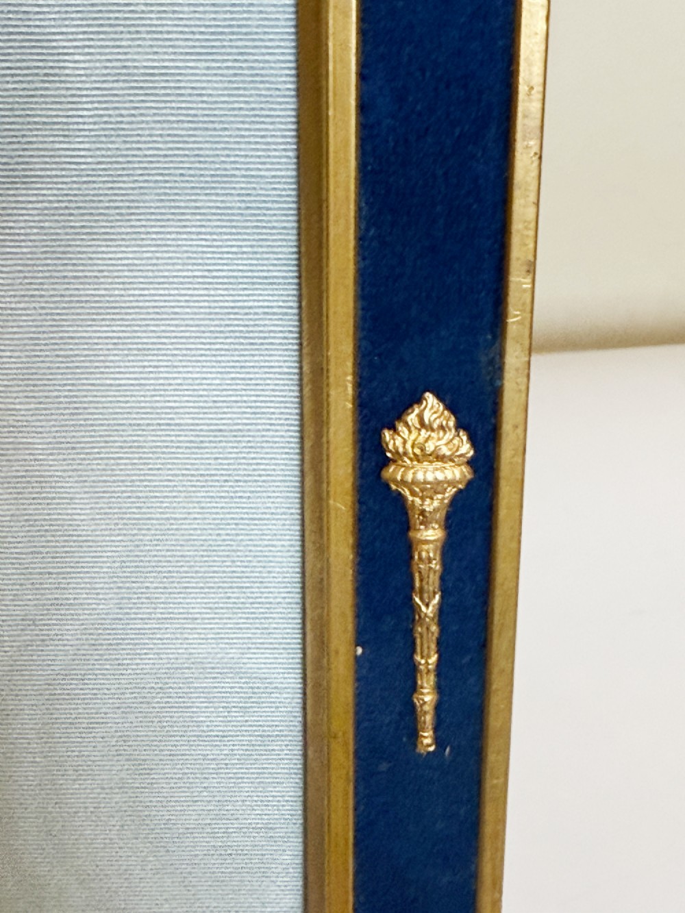 A rectangular gilt bronze picture frame featuring a blue velvet border, ornate corner mounts, and an elaborate floral crest at the top. View V