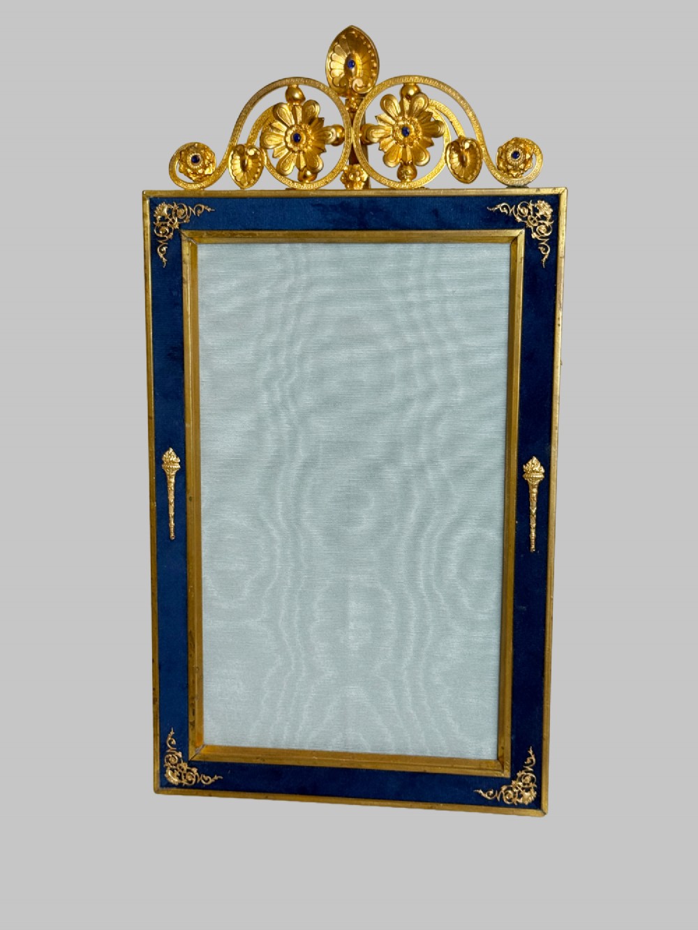 A rectangular gilt bronze picture frame featuring a blue velvet border, ornate corner mounts, and an elaborate floral crest at the top. View X