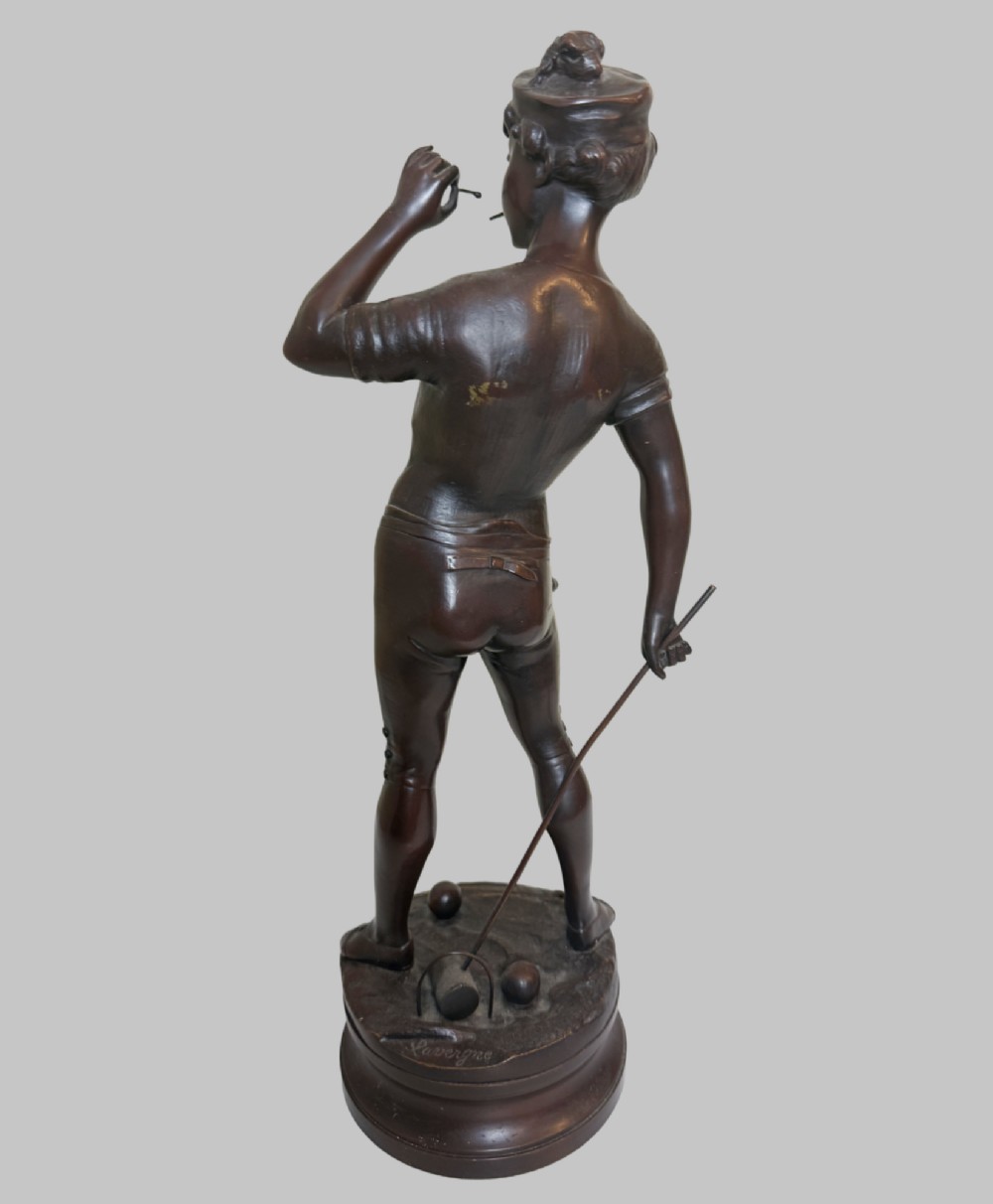 A dark patinated bronze statue of a young man smoking a cigarette while holding a croquet mallet on a round base with croquet balls. View II