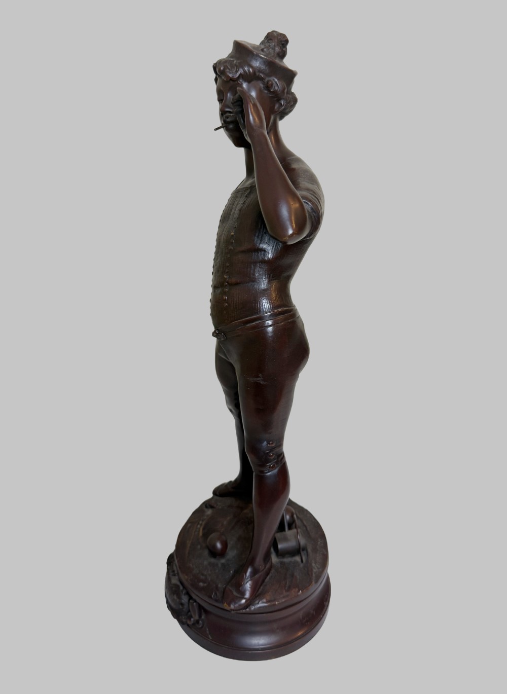 A dark patinated bronze statue of a young man smoking a cigarette while holding a croquet mallet on a round base with croquet balls. View III