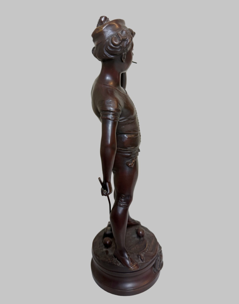 A dark patinated bronze statue of a young man smoking a cigarette while holding a croquet mallet on a round base with croquet balls. View IV